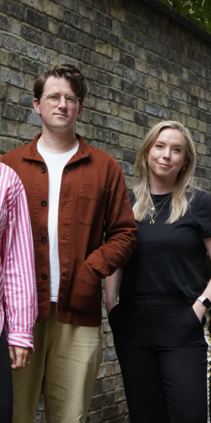 A group of six people stands in a narrow alleyway between two brick walls, posing for the photo. From left to right: 1. Simon Jenkins in a light blue shirt with rolled-up sleeves and dark green pants, standing with his hand in his pocket. 2. Becky Marsh with long blonde hair wearing a pink and white striped shirt and black pants, smiling. 3. Will Parrish with brown hair wearing glasses, a white t-shirt, and a brown jacket over beige pants. 4. Lindsay Turner with blonde hair wearing a black t-shirt and black pants, with her hands in her pockets. 5. Kim Jamieson stands with blonde hair wearing a green jumpsuit, also with her hands in her pockets. 6. James Shoreland leaning against a black lamppost. The group appears relaxed and friendly, with a backdrop of an urban brick alley and some greenery above.