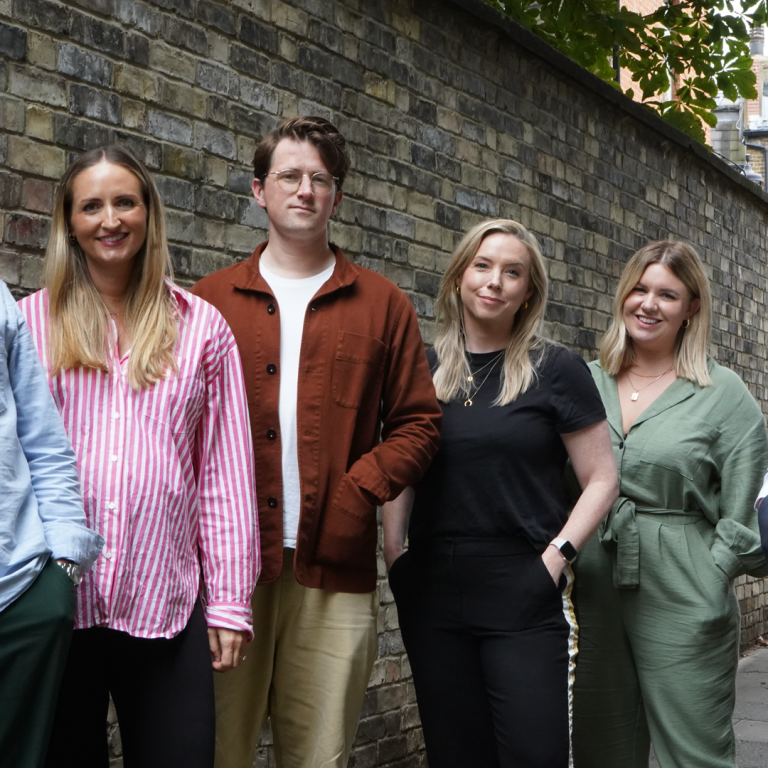 A group of six people stands in a narrow alleyway between two brick walls, posing for the photo. From left to right: 1. Simon Jenkins in a light blue shirt with rolled-up sleeves and dark green pants, standing with his hand in his pocket. 2. Becky Marsh with long blonde hair wearing a pink and white striped shirt and black pants, smiling. 3. Will Parrish with brown hair wearing glasses, a white t-shirt, and a brown jacket over beige pants. 4. Lindsay Turner with blonde hair wearing a black t-shirt and black pants, with her hands in her pockets. 5. Kim Jamieson stands with blonde hair wearing a green jumpsuit, also with her hands in her pockets. 6. James Shoreland leaning against a black lamppost. The group appears relaxed and friendly, with a backdrop of an urban brick alley and some greenery above.